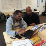 Two men working on a laptop together in a classroom
