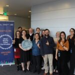 A group of City employees smiling and holding up one finger for "One Seattle." They are standing next to a station and holding an award showing that Seattle, WA earned a What Works Cities Certification.
