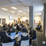 A crowd of hackathon attendees, sitting in groups around their tables, listen to a City staff member giving instructions.