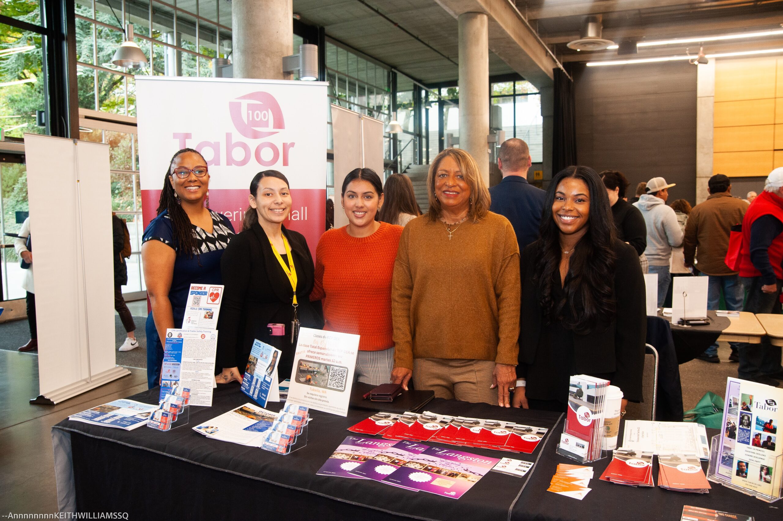 A group of five women smile for a photo behind a table full of pamphlets and hand-outs. A sign behind them has the Tabor 100 logo