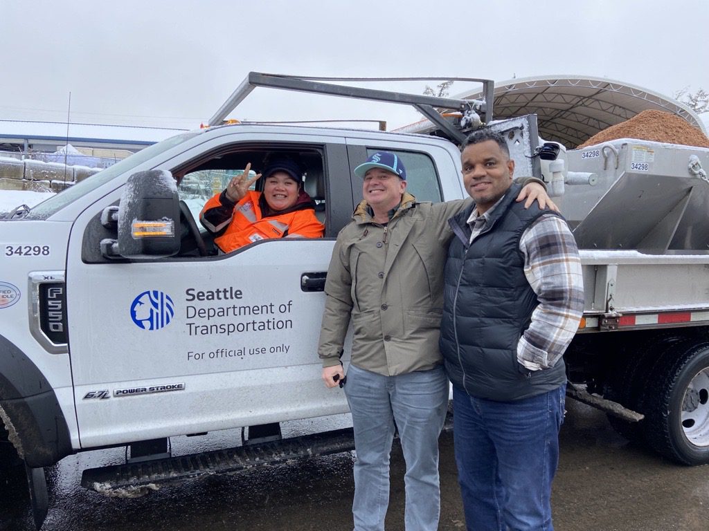 A driver flashes a peace sign from the drivers seat of an SDOT snow plow, with two other SDOT employees smiling for a photo next to the vehicle