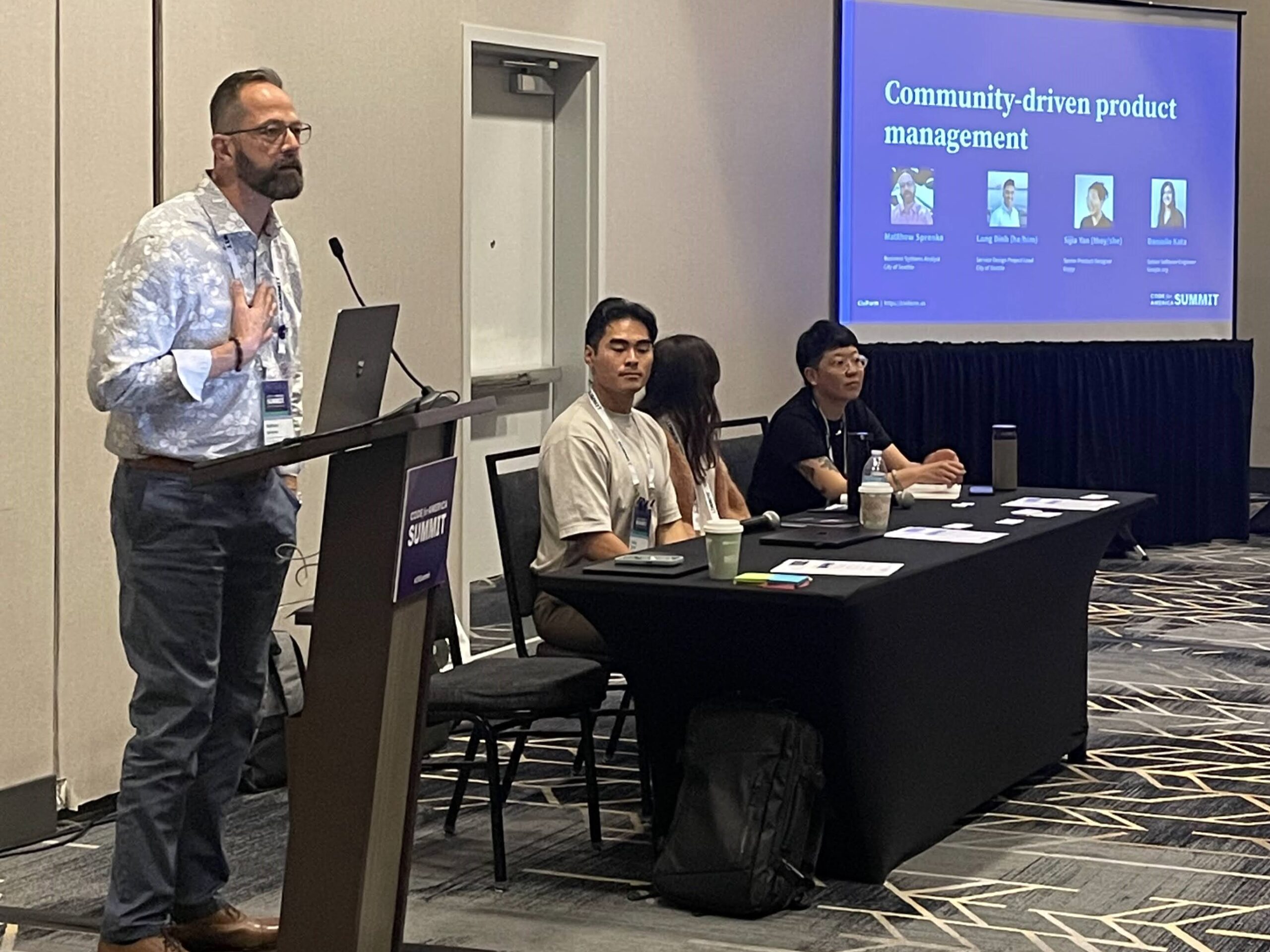 A man speaking at a podium next to a table of three other panelists, with a screen in the background that says "Community-driven product management"