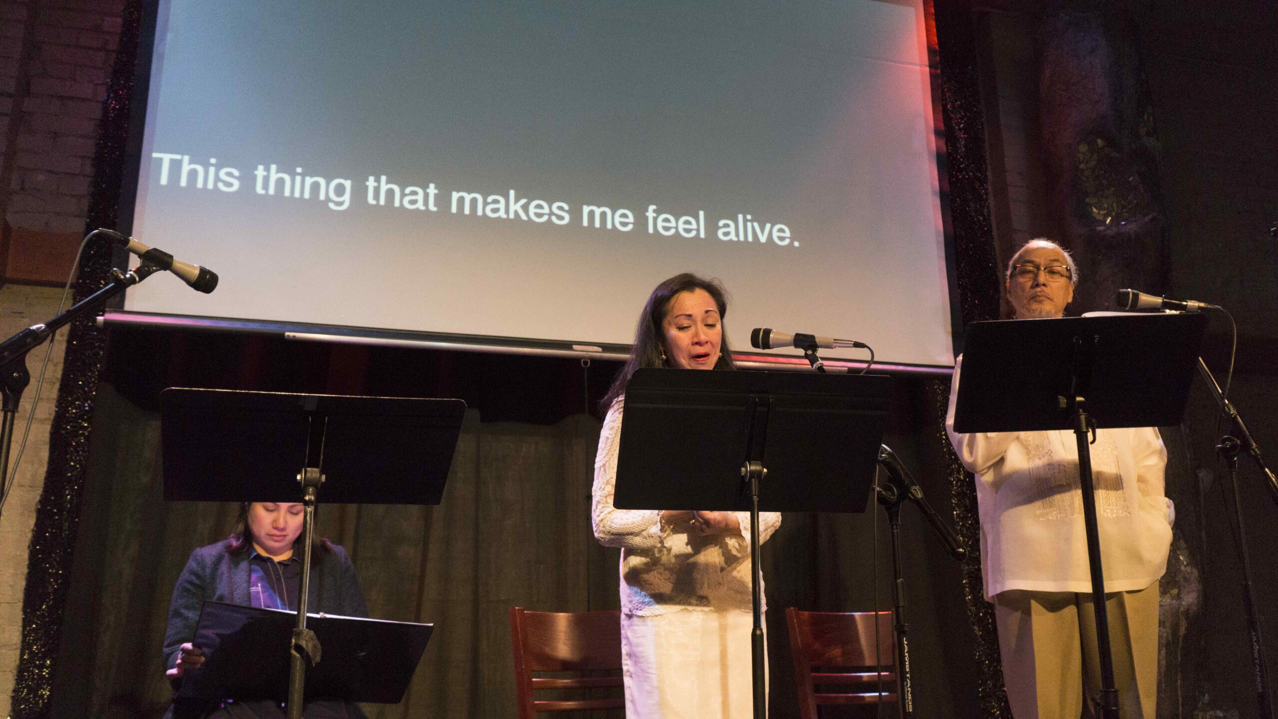 A woman reading aloud on a stage, with a screen behind her displaying the words "This thing that makes me feel alive."