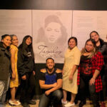 A group of 7 actors pose for a photo together in front of a poster for their show