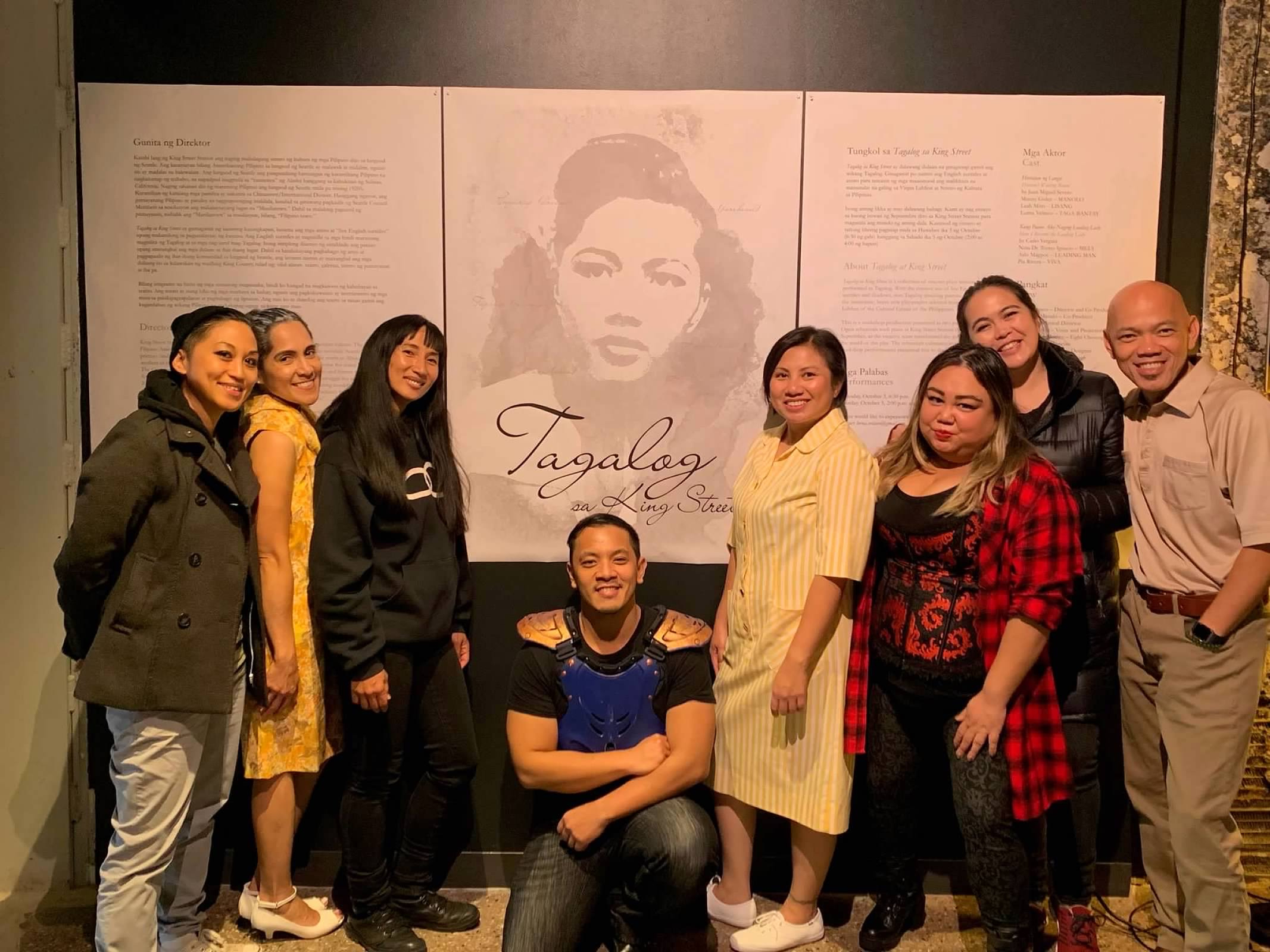 A group of 7 actors pose for a photo together in front of a poster for their show