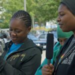 Two teenagers, one holding a camera while the other speaks into a mircophone