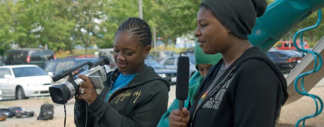 Two teenagers, one holding a camera while the other speaks into a mircophone