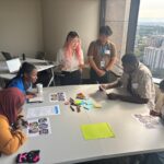 A group of four teenagers sitting around a table working on an activity together. Two more teenagers, Ada and Tony, stand next to the table facilitating the group activitity