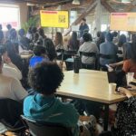 A large group of people sitting at tables listening to a presenter at the Youth Connector Hackathon