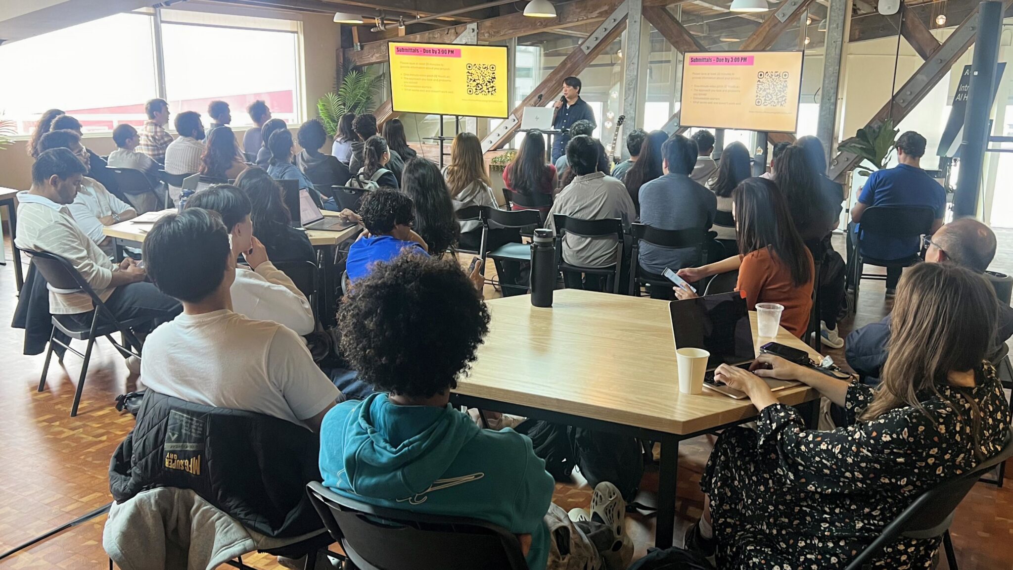 A large group of people sitting at tables listening to a presenter at the Youth Connector Hackathon